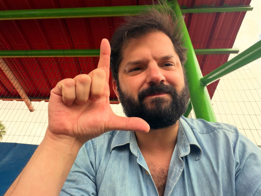 Homem de barba e cabelo escuro faz o gesto do L com a mão.